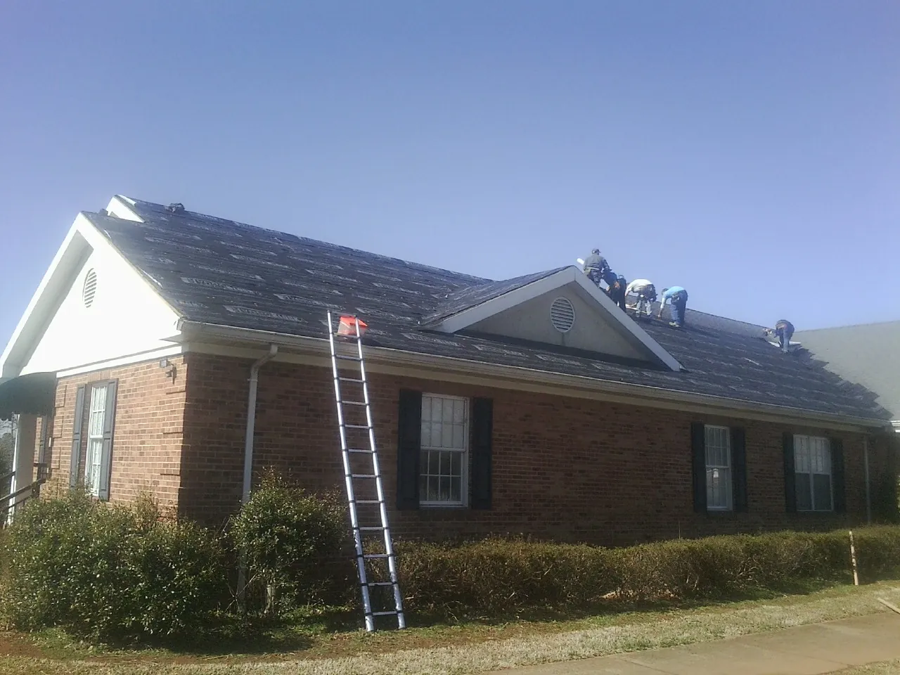 Crew tearing shingles off a brick ranch roof with a ladder set — Metal Roof Repair work by our crew in Eskridge