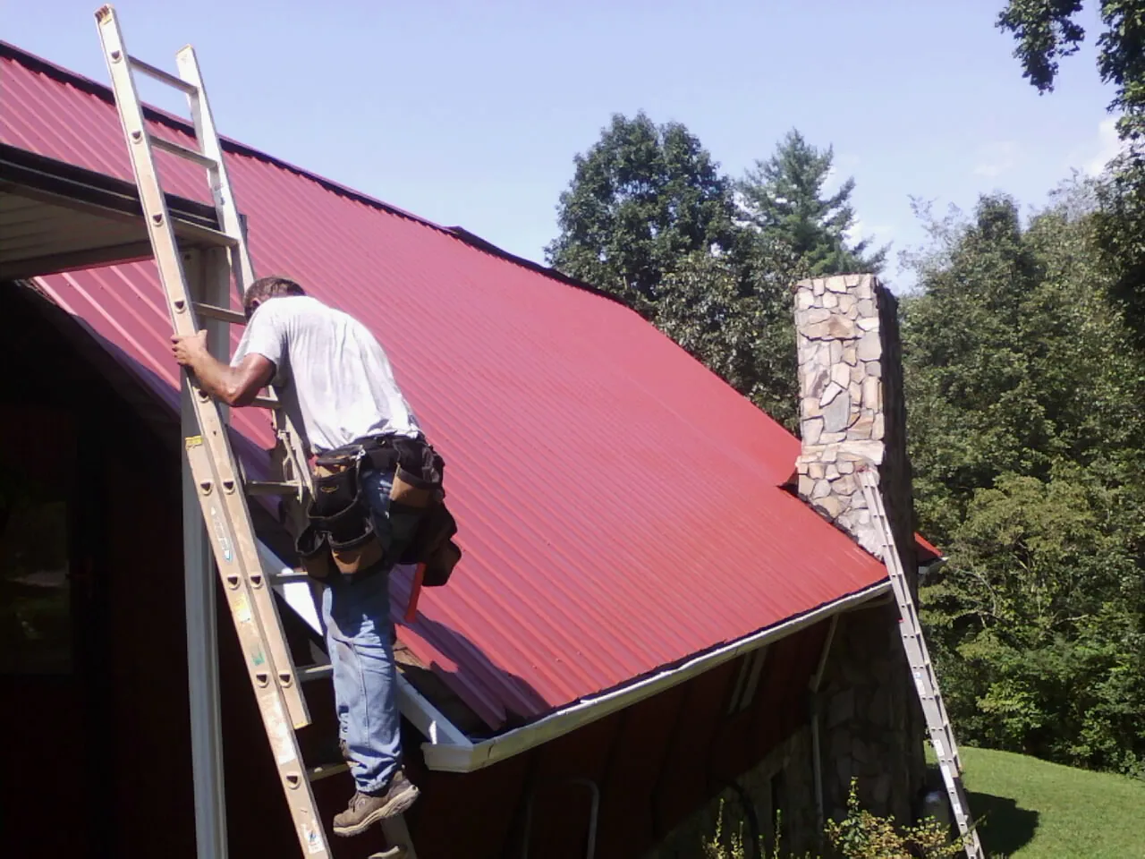 Roofer on a ladder beside a red metal roof and stone chimney — Soffit & Fascia Repair by Roofing Brothers in Eskridge